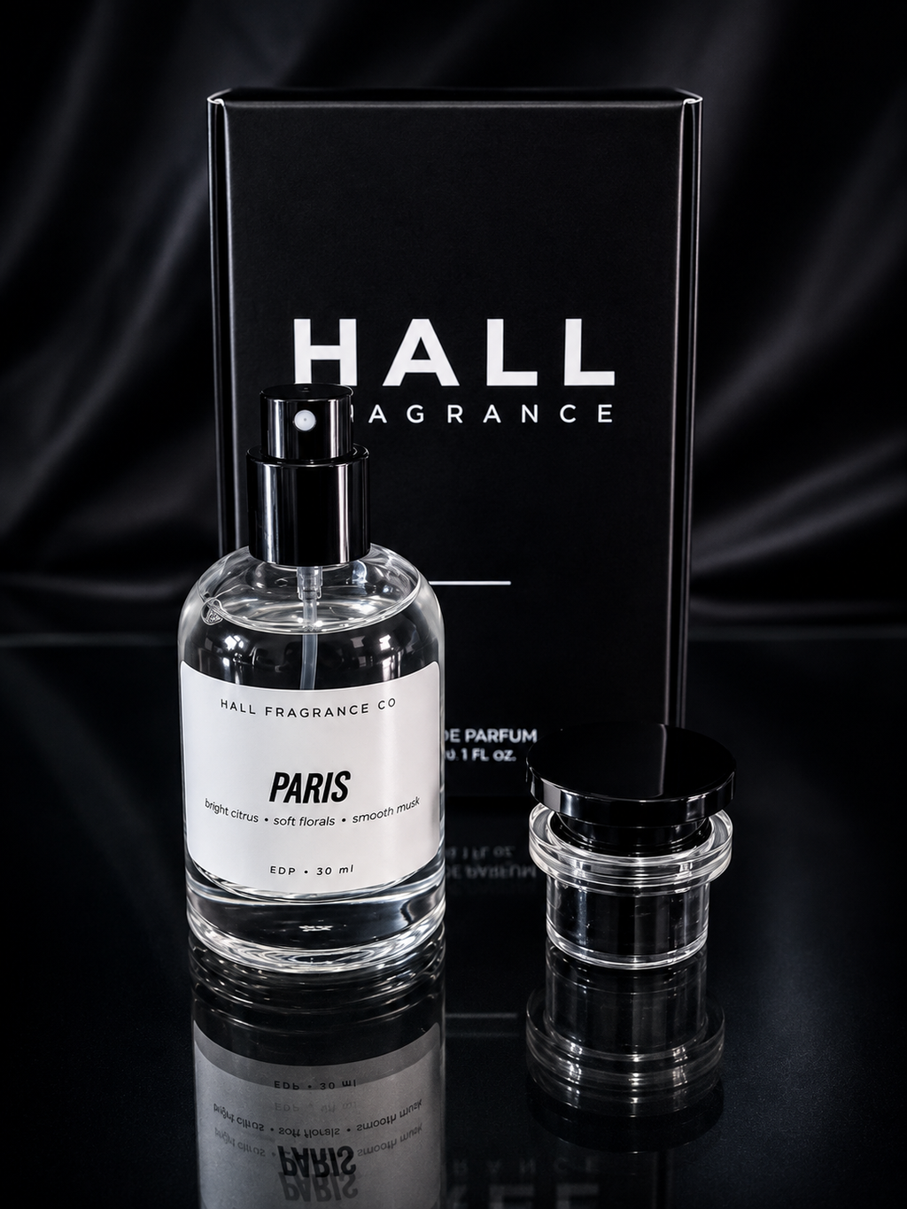 A clear glass perfume bottle labeled PARIS by Hall Fragrance Co stands in front of a tall black product box with large white HALL FRAGRANCE text, with a small clear jar with a black lid beside it on a dark reflective surface.