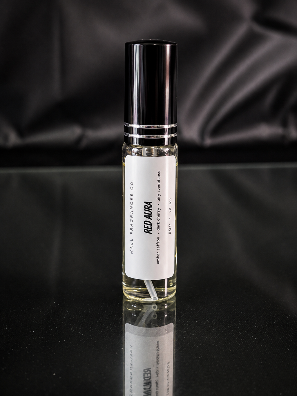 A 10 ml rollerball bottle of Hall Fragrance Co. 'Red Aurora' perfume, featuring a clear glass bottle with pale yellow liquid, a white label, and a glossy black cap, set against a dark draped satin background.